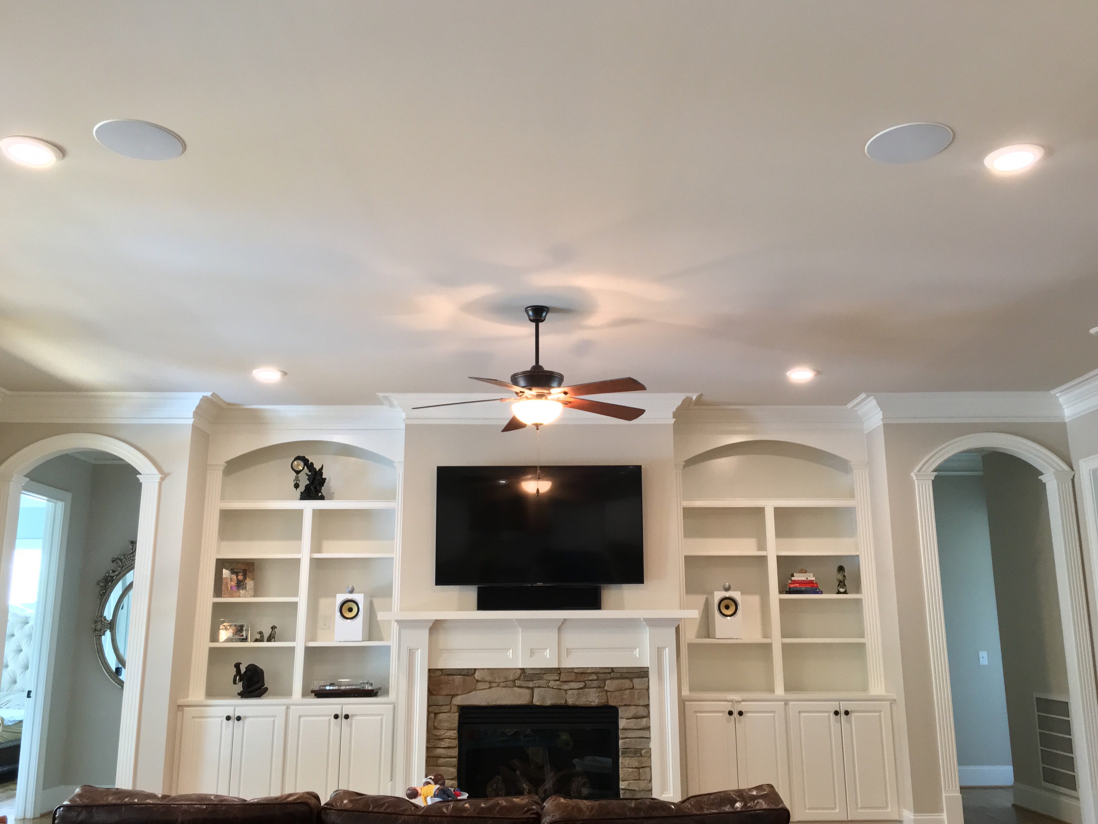 A living room with ceiling fan, recessed lights, mounted TV above a fireplace, and white built-in shelves.