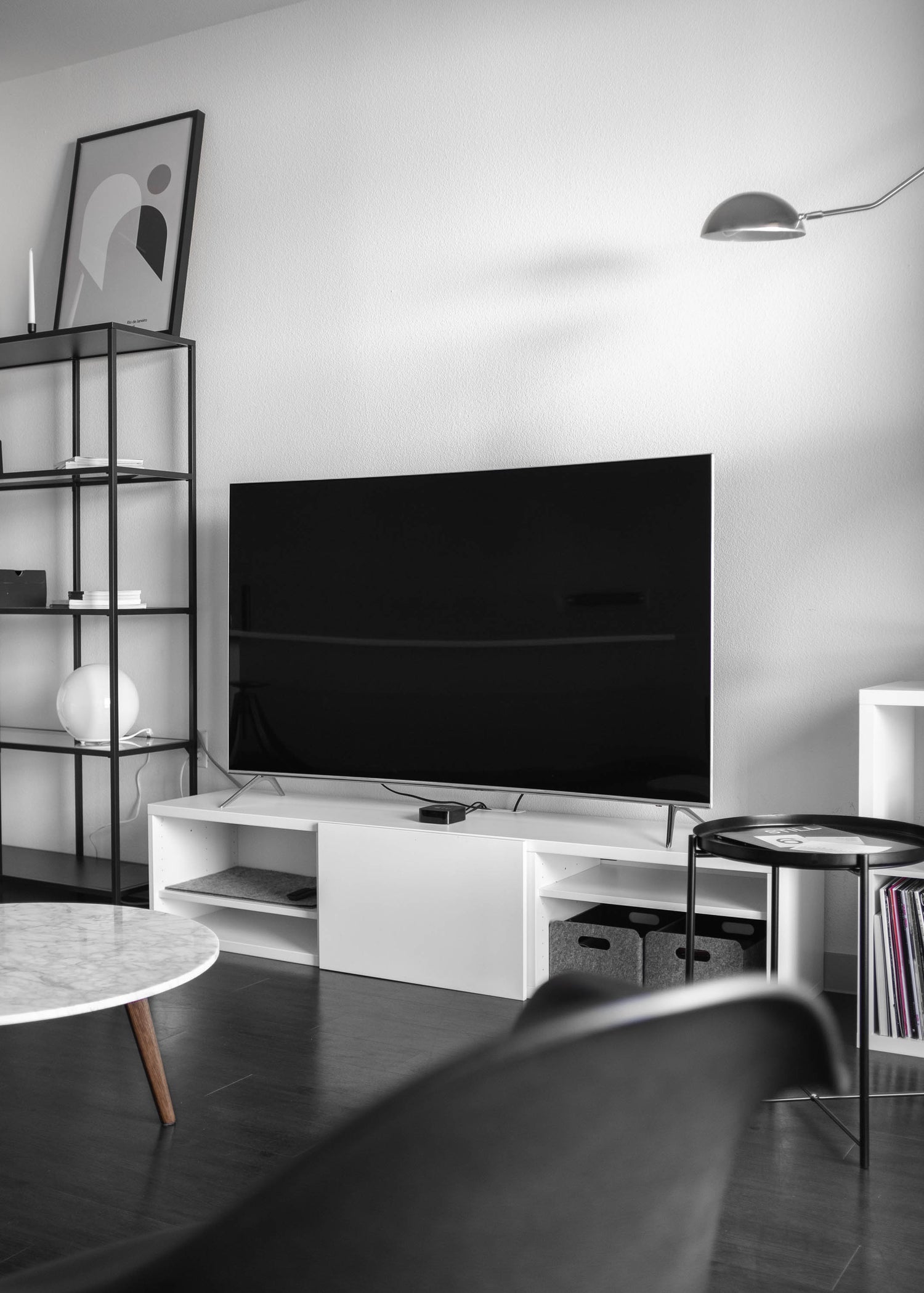 A monochromatic living room featuring a flat-screen TV on a white stand, with a minimalist metal shelving unit and modern decor in the background.