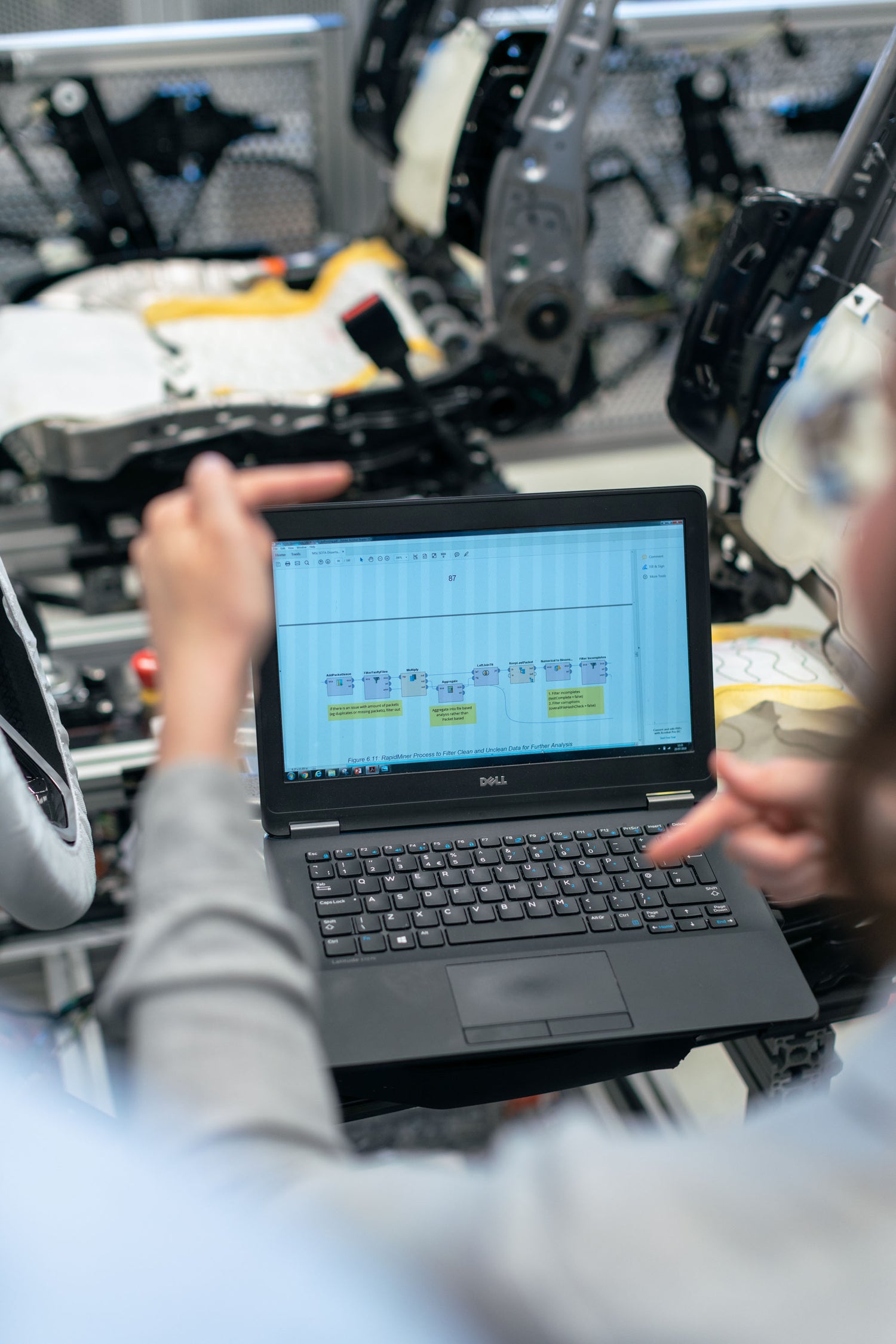 Over-the-shoulder view of an engineer working on a laptop with a vehicle assembly diagram on the screen, car parts and components visible in the background.