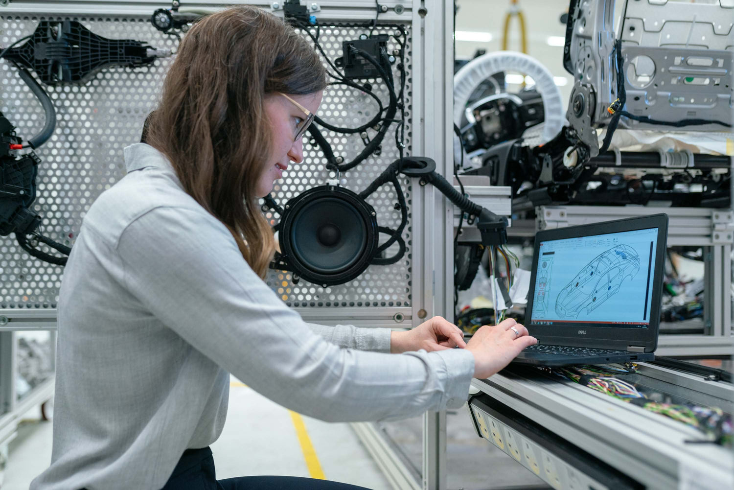 A female engineer working with a laptop, the screen displaying a car design diagram, with automotive parts mounted on the wall behind her.